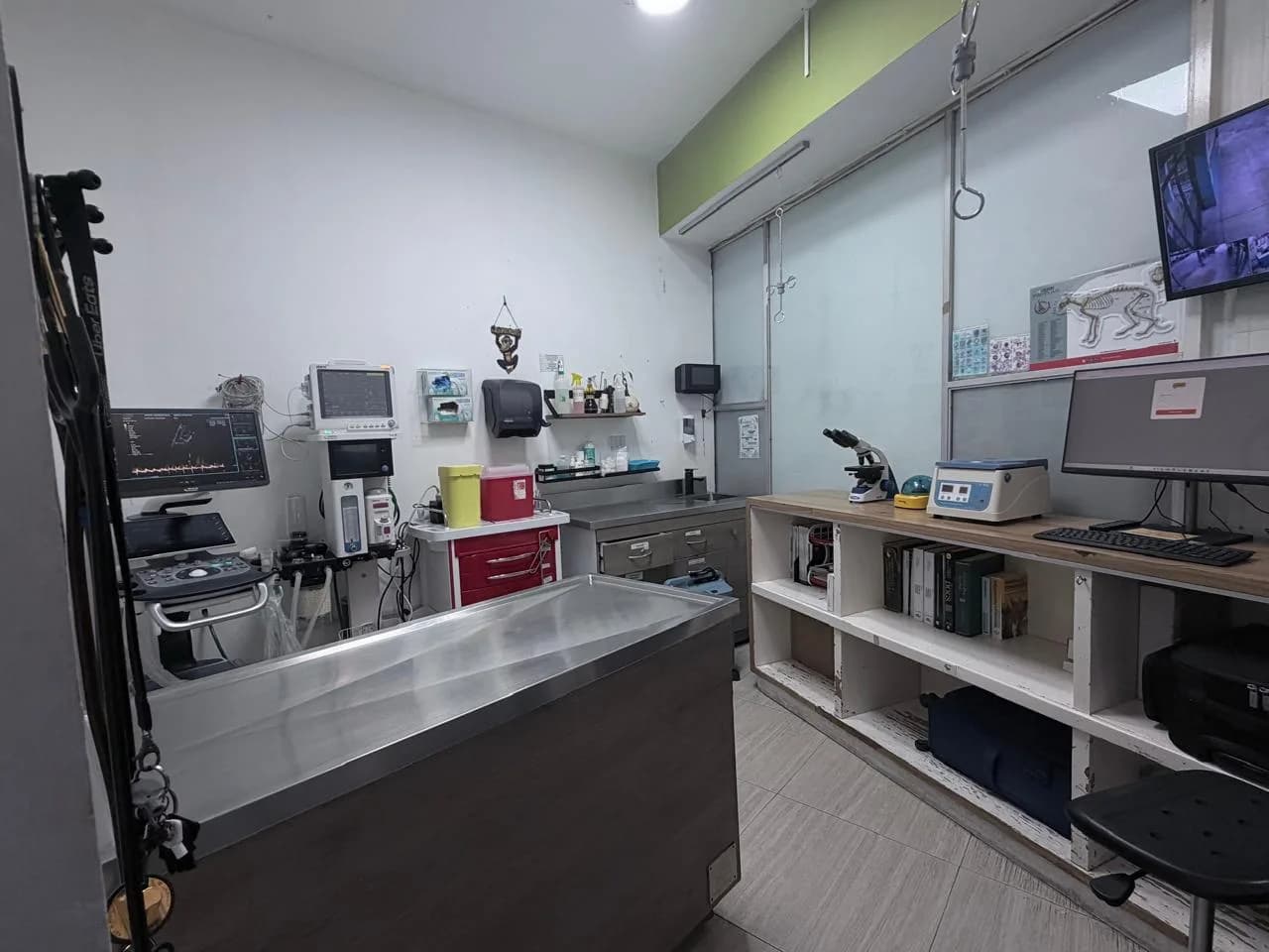 Consultorio con ultrasonido, monitor de signos vitales, microscopio y centrífuga junto a la mesa de exploración de acero