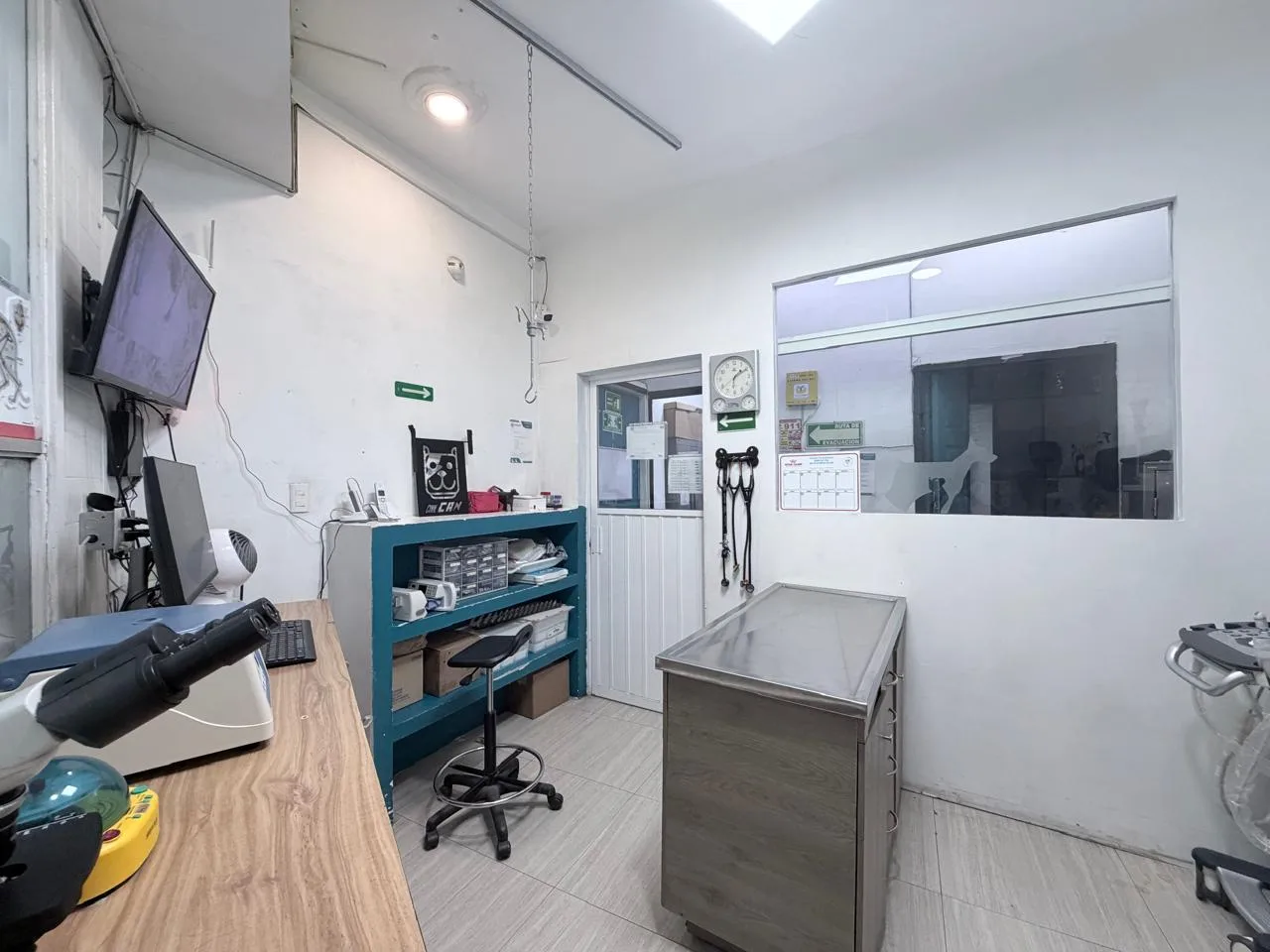 Consultorio con microscopio en la barra, estetoscopios en la pared y mesa de exploración de acero junto a la ventana interior