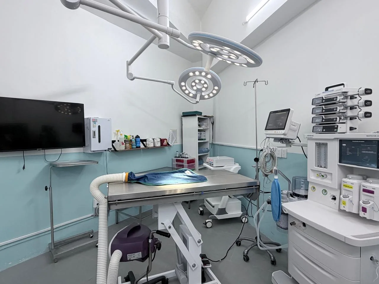 Quirófano con mesa de cirugía de acero, dos lámparas quirúrgicas, monitor de signos vitales y pantalla en la pared
