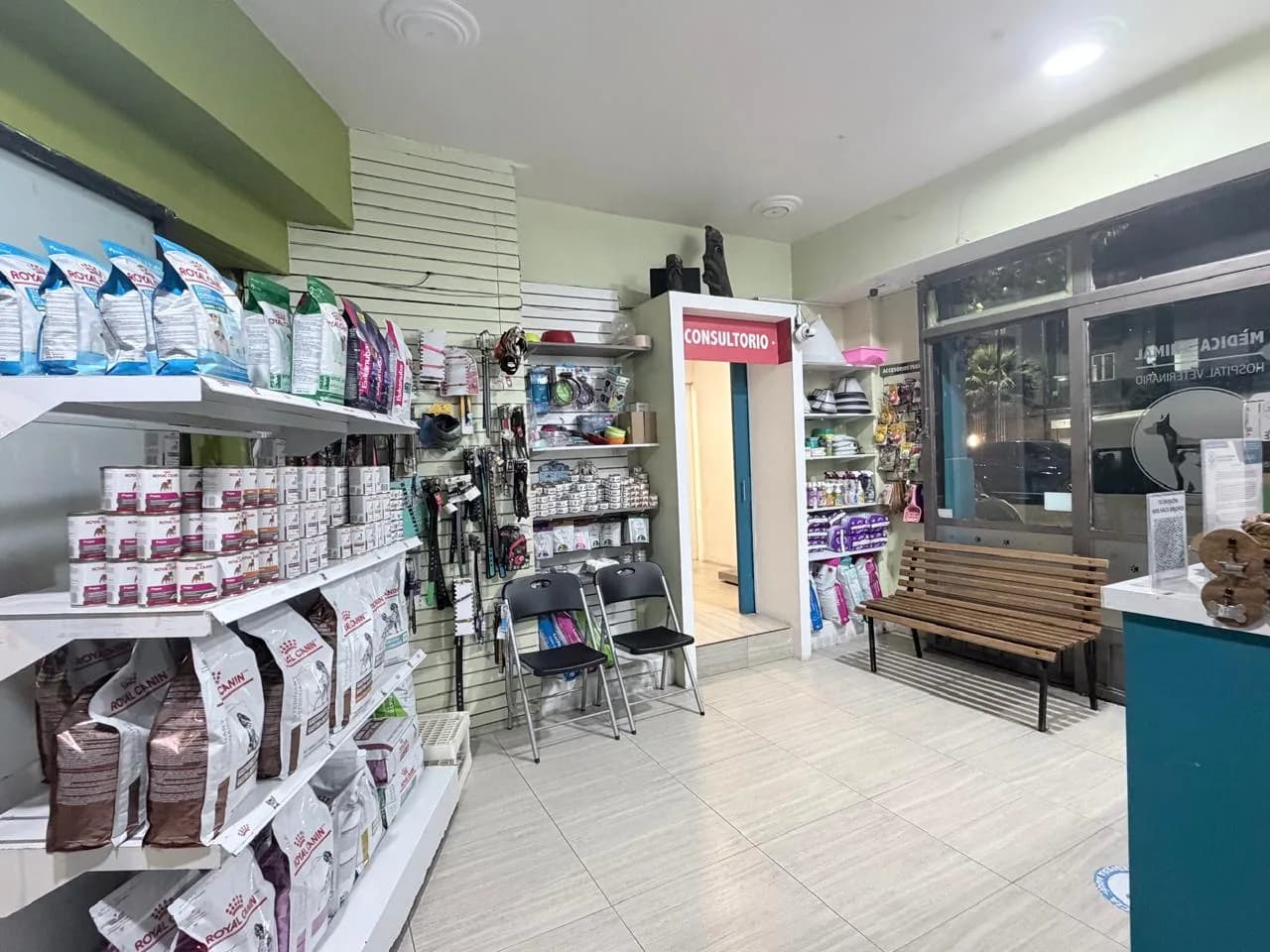 Tienda y zona de espera con anaqueles de alimento, sillas, banca de madera junto al ventanal y la puerta del consultorio al fondo